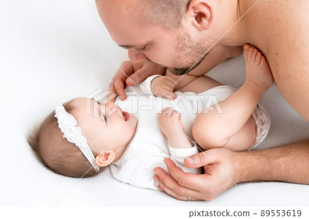 A little innocent baby girl smiles sweetly and tenderly looking into Dad's eyes. An infant with his father in a snow-white bed. Fatherhood and upbringing in love and harmony A little innocent baby girl smiles sweetly and tenderly looking into Dad's eyes. An infant with his father in a snow-white bed. Fatherhood and upbringing in love and harmony 89553619