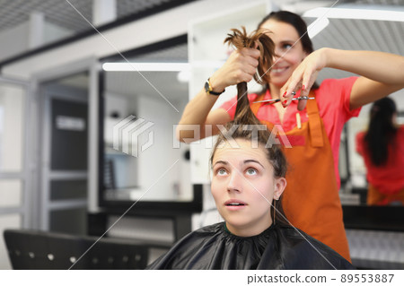 Hairdresser going to cut lock of long hair with scissors tool, female client scared in masters chair 89553887