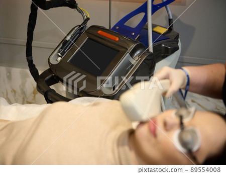 Focus on modern laser apparatus in medical spa clinic with blurred Caucasian woman getting hair removal treatment on her face in the foreground 89554008