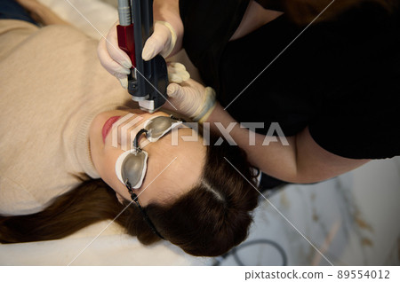 Young pretty woman wearing UV protective goggles, lying down on daybed while receiving hair removal laser procedure on her face in a wellness spa clinic Young pretty woman wearing UV protective goggles, lying down on daybed while receiving hair removal laser procedure on her face in a wellness spa clinic 89554012