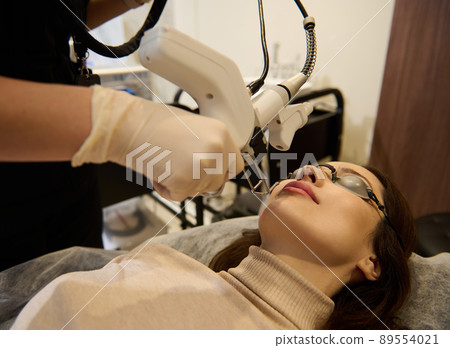Close-up of a Caucasian middle-aged woman getting facial laser polishing and rejuvenation face procedure, cosmetologist using hardware apparatus Close-up of a Caucasian middle-aged woman getting facial laser polishing and rejuvenation face procedure, cosmetologist using hardware apparatus 89554021