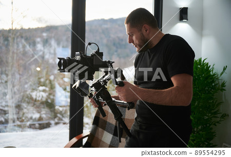 Videographer man shooting footage, using camera mounted on gimbal stabilizer equipment. Panoramic windows with winter landscape on background. 89554295
