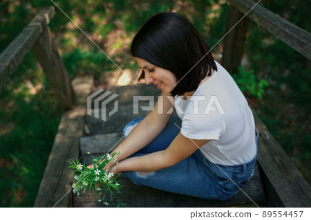 A young beautiful woman sat down to rest on an old wooden bridge in the forest and looks at her collected bouquet of wild flowers. 89554457