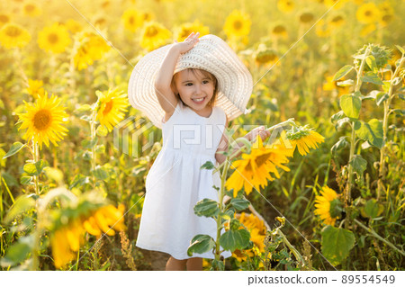 A cheerful cute girl in a hat with a large brim is standing in a field of sunflowers at sunset. Happy childhood. A cheerful cute girl in a hat with a large brim is standing in a field of sunflowers at sunset. Happy childhood. 89554549