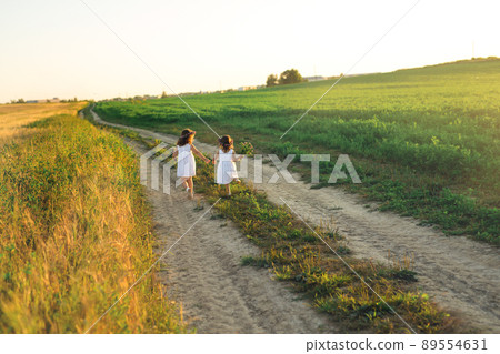 Two girls run along a country road. The road between the wheat and clover fields. Landscape at sunset. Girls in white dresses and with a wreath on their heads. 89554631
