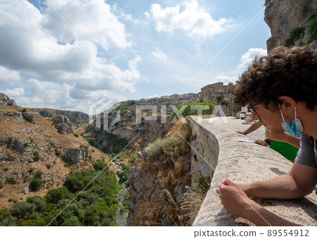 Matera, Basilicata, Italy. August 2021. A young tourist has dropped his mask enjoying the view of the narrow valley of the Gravina river. The village in the background. Matera, Basilicata, Italy. August 2021. A young tourist has dropped his mask enjoying the view of the narrow valley of the Gravina river. The village in the background. 89554912