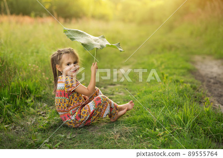 It's starting to rain. The girl is hiding under a large burdock. Cute baby at sunset. Girl hold large burdock leaf like umbrella. 89555764