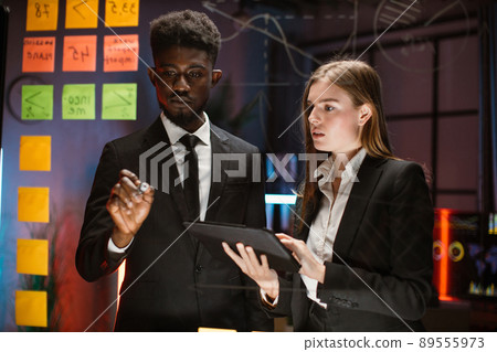 Young diverse multiethnic coworkers, standing near the glass board, writing and using tablet pc Young diverse multiethnic coworkers, standing near the glass board, writing and using tablet pc 89555973