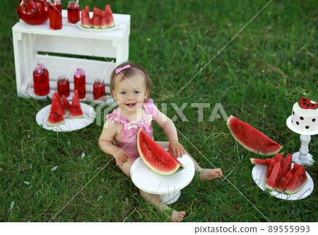 A happy girl looks up at the camera sitting on the lawn with a large piece of sweet juicy watermelon. National Watermelon Day. A festive picnic on a child's birthday 89555993