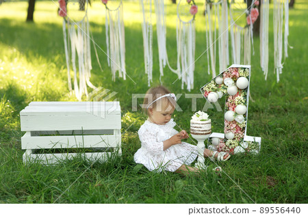 A girl in a smart white dress is sitting on the grass and looking at her hands soiled in cream. Birthday cake for the first birthday and decorations with ribbons on trees in the garden A girl in a smart white dress is sitting on the grass and looking at her hands soiled in cream. Birthday cake for the first birthday and decorations with ribbons on trees in the garden 89556440