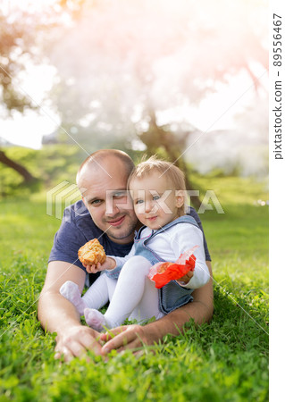 A little girl is eating a pie in her father's arms and laughing merrily. The relationship between father and daughter. Picnic in the park at sunset. A loving father. Happy family. Childhood in harmony A little girl is eating a pie in her father's arms and laughing merrily. The relationship between father and daughter. Picnic in the park at sunset. A loving father. Happy family. Childhood in harmony 89556467