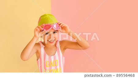Funny joy girl in striped swimsuit and a pool cap, puts on swimming glasses, isolated on an pink and yellow studio background. Learning to swim in the pool from an early age. Happy baby Funny joy girl in striped swimsuit and a pool cap, puts on swimming glasses, isolated on an pink and yellow studio background. Learning to swim in the pool from an early age. Happy baby 89556468