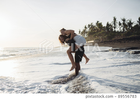 a man walks along the water's edge in the ocean and carries a woman on her shoulders against the backdrop of coastal palm trees 89556783