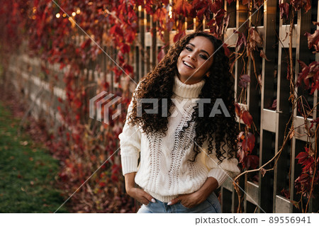 a curly-haired brunette with a charming white-toothed smile clad in a white sweater and blue jeans leaned against the red ivy wall a curly-haired brunette with a charming white-toothed smile clad in a white sweater and blue jeans leaned against the red ivy wall 89556941