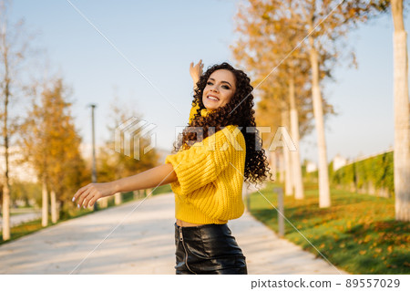 A girl in a bright sweater with long black curly hair dances in the alley of the park surrounded by autumn trees. 89557029