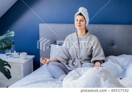 Woman in pajamas and bathrobe with eyes patches and towel on head practicing meditation sitting on bed. Home Beauty routine and morning self-care, self-healing, mindfulness. Mental health practice. 89557592