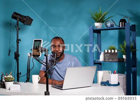 Smiling influencer interacting with audience in front of recording smartphone during online live show using laptop. Content creator talking with fanbase looking at live video podcast setup. Smiling influencer interacting with audience in front of recording smartphone during online live show using laptop. Content creator talking with fanbase looking at live video podcast setup. 89568935