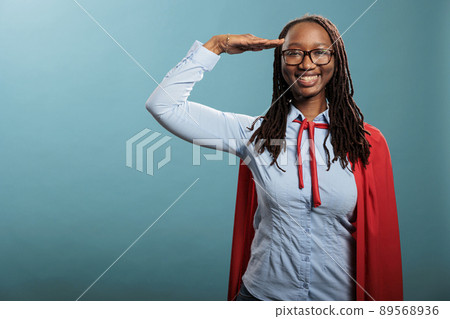Patriotic justice defender saluting with respect while standing on blue background while smiling at camera. Brave and proud happy superhero woman wearing mighty hero cloak while making soldier salute. 89568936
