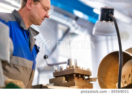 Elderly turner of 50-55 years old in overalls works at lathe in production shop. Processing of metal Elderly turner of 50-55 years old in overalls works at lathe in production shop. Processing of metal 89569031