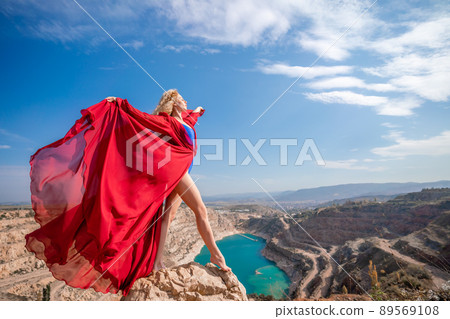 Side view of a beautiful sensual woman in a red long dress posing on a rock high above the lake in the afternoon. Against the background of the blue sky and the lake in the form of a heart 89569108