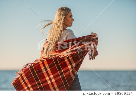 Attractive blonde Caucasian woman enjoying time on the beach at sunset, walking in a blanket and looking to the side, with the sunset sky and sea in the background. Beach vacation 89569134