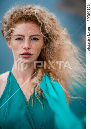 Outdoor portrait of a young beautiful natural redhead girl with freckles, long curly hair, in an emerald dress, posing against the background of the sea. Outdoor portrait of a young beautiful natural redhead girl with freckles, long curly hair, in an emerald dress, posing against the background of the sea. 89569176
