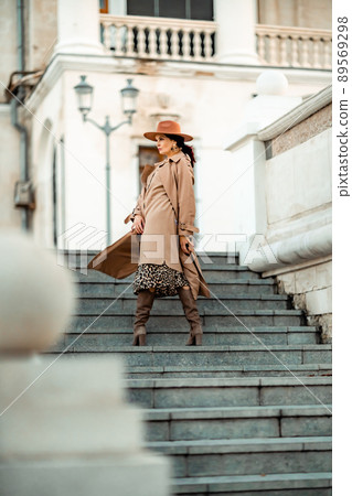 Outdoor fashion portrait of young elegant fashionable brunette woman, model in stylish hat, choker and light raincoat posing at sunset in European city. Outdoor fashion portrait of young elegant fashionable brunette woman, model in stylish hat, choker and light raincoat posing at sunset in European city. 89569298