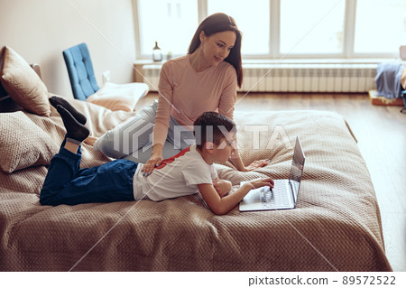 Beautiful young Caucasian mother sitting on bed and watching small son using laptop. Beautiful young Caucasian mother sitting on bed and watching small son using laptop. 89572522