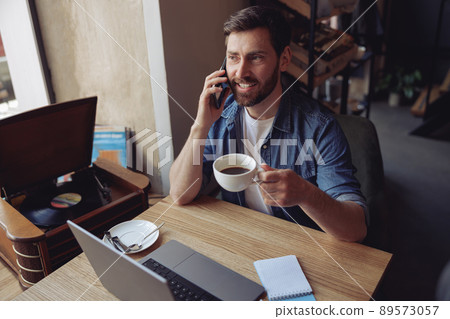 Attractive Caucasian man talking on mobile phone and drinking coffee in cafe. Business chat. Attractive Caucasian man talking on mobile phone and drinking coffee in cafe. Business chat. 89573057
