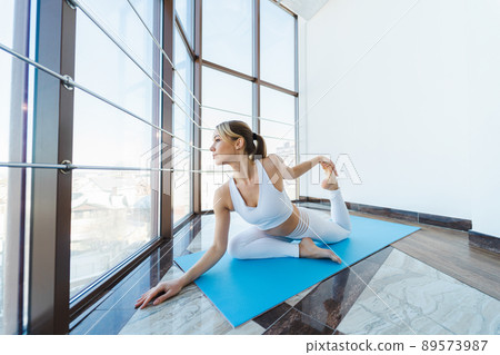 Pretty blonde woman doing yoga exercise while looking at the city through the panoramic windows 89573987