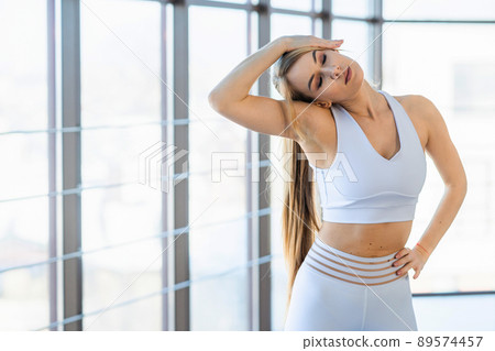 Beautiful girl in white sportswear warms up against the background of panoramic windows 89574457