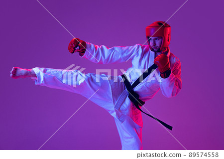 Studio shot of male taekwondo fighter in sports uniform in action isolated over purple background in neon light. Concept of sport, workout, competition, ad Studio shot of male taekwondo fighter in sports uniform in action isolated over purple background in neon light. Concept of sport, workout, competition, ad 89574558