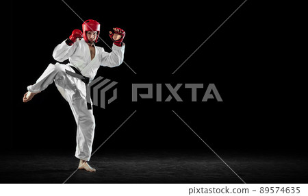 Male taekwondo fighter in white dobok, helmet and gloves training isolated over dark background. Concept of sport, workout, competition, ad Male taekwondo fighter in white dobok, helmet and gloves training isolated over dark background. Concept of sport, workout, competition, ad 89574635