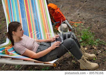 Caucasian woman working on laptop while sitting in a hammock in the forest. Girl uses a wireless computer on a hike. Caucasian woman working on laptop while sitting in a hammock in the forest. Girl uses a wireless computer on a hike. 89576166