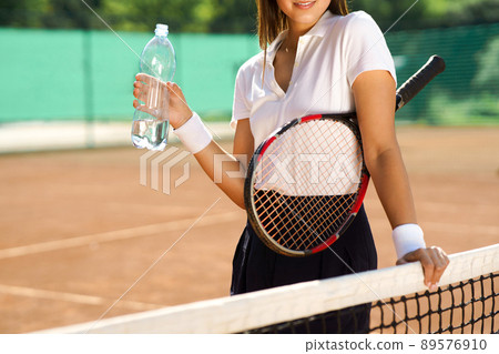 Young woman tennis player with a bottle of water stands on the court Young woman tennis player with a bottle of water stands on the court 89576910