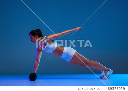 Portrait of young sportive woman standing on plank with dumbbell isolated over blue studio backgtound in neon light 89578959