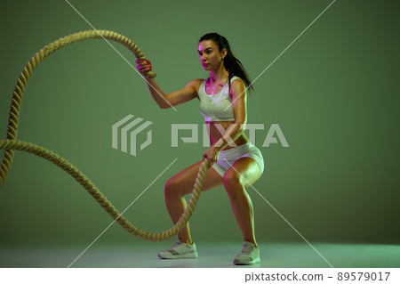 Dynamic portrait of young sportive woman training, doing exercises with rope isolated over green studio background in neon light Dynamic portrait of young sportive woman training, doing exercises with rope isolated over green studio background in neon light 89579017