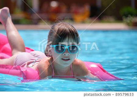Young child girl relaxing on summer sun swimming on inflatable air mattress in swimming pool during tropical vacations. Summertime activities concept Young child girl relaxing on summer sun swimming on inflatable air mattress in swimming pool during tropical vacations. Summertime activities concept 89579633