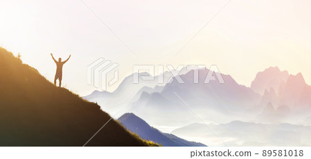 Man on peak of mountain. Emotional scene. Young man with backpack standing with raised hands on top of a mountain and enjoying mountain view. Hiker on the mountain top. Sport and active life concept. 89581018