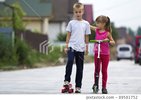 Cute young blond children, brother and sister, girl in pink clothing on scooter and handsome boy on skateboard playing together on sunny street blurred background. Children games and fun concept. Cute young blond children, brother and sister, girl in pink clothing on scooter and handsome boy on skateboard playing together on sunny street blurred background. Children games and fun concept. 89581752