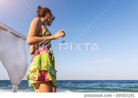 Back view young stylish adult woman holding cold alcohol refresh beverage glass looking at ocean shore at summer seaside vacation. Female person treat relax holiday at exotic resort hotel beach 89583191