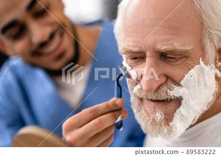 Joyful caregiver shaving an aged man with a razor Joyful caregiver shaving an aged man with a razor 89583222