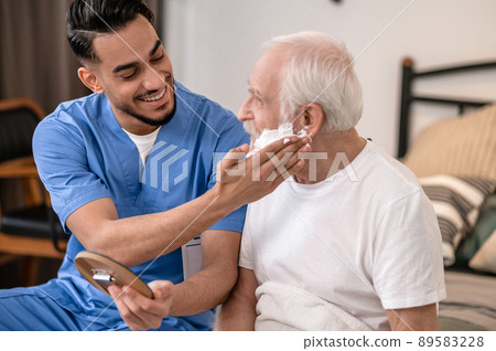 Joyful young volunteer preparing an old man for a shave Joyful young volunteer preparing an old man for a shave 89583228