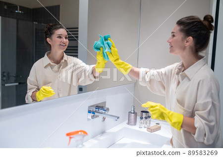 Cute young woman cleaning mirror in the bathroom Cute young woman cleaning mirror in the bathroom 89583269