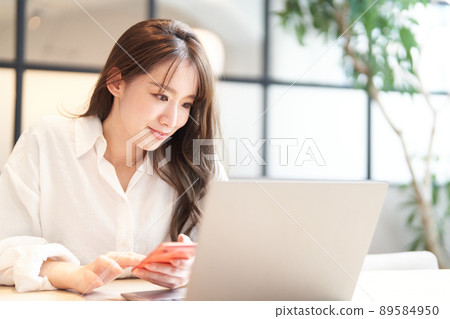 一名年輕女子一邊看筆記本電腦的屏幕一邊使用智能手機 89584950