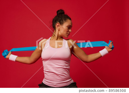 African American muscular woman doing stretching exercises on arms using elastic fitness band against red background with copy ad space African American muscular woman doing stretching exercises on arms using elastic fitness band against red background with copy ad space 89586308