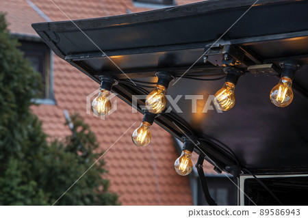 Blurred background, backyard illumination, light in the garden, electric lanterns with round diffuser. Lamp garland of light bulbs, illumination Blurred background, backyard illumination, light in the garden, electric lanterns with round diffuser. Lamp garland of light bulbs, illumination 89586943