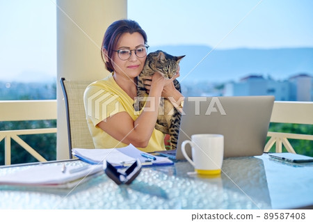 Middle-aged woman working at home with a cat in her arms. 89587408