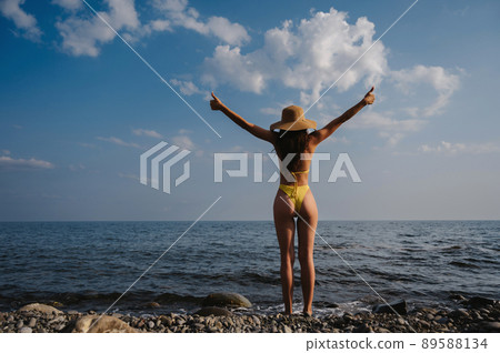 girl with a sexy booty with a straw hat in a swimsuit raises her hands up while standing with her back on beach by sea 89588134