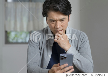 A middle-aged man who thinks while looking at the screen of a smartphone A middle-aged man who thinks while looking at the screen of a smartphone 89599511